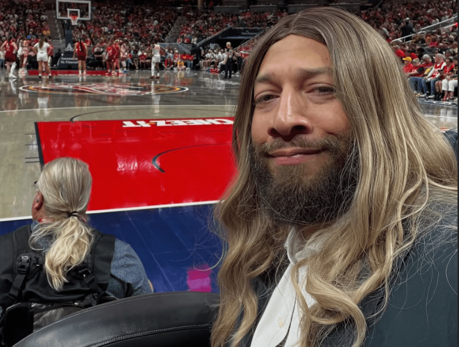 A bearded man with long blond hair sits in a basketball arena, with a large red "Adidas" logo visible in the background.
